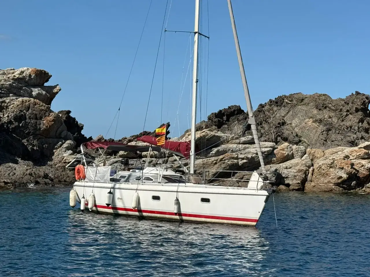 Voilier blanc à la dérive, ancré près de rochers escarpés. Un drapeau catalan flotte au mât. Étape 32i avis.
