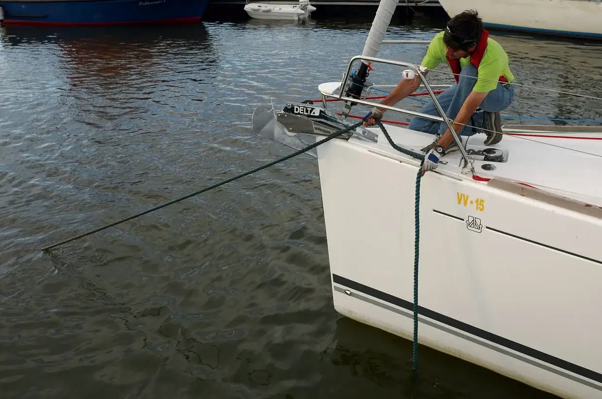 Un homme en gilet de sauvetage ajuste l'amarrage du bateau avec une pendille. L'ancre Delta est visible &agrave; l'avant.