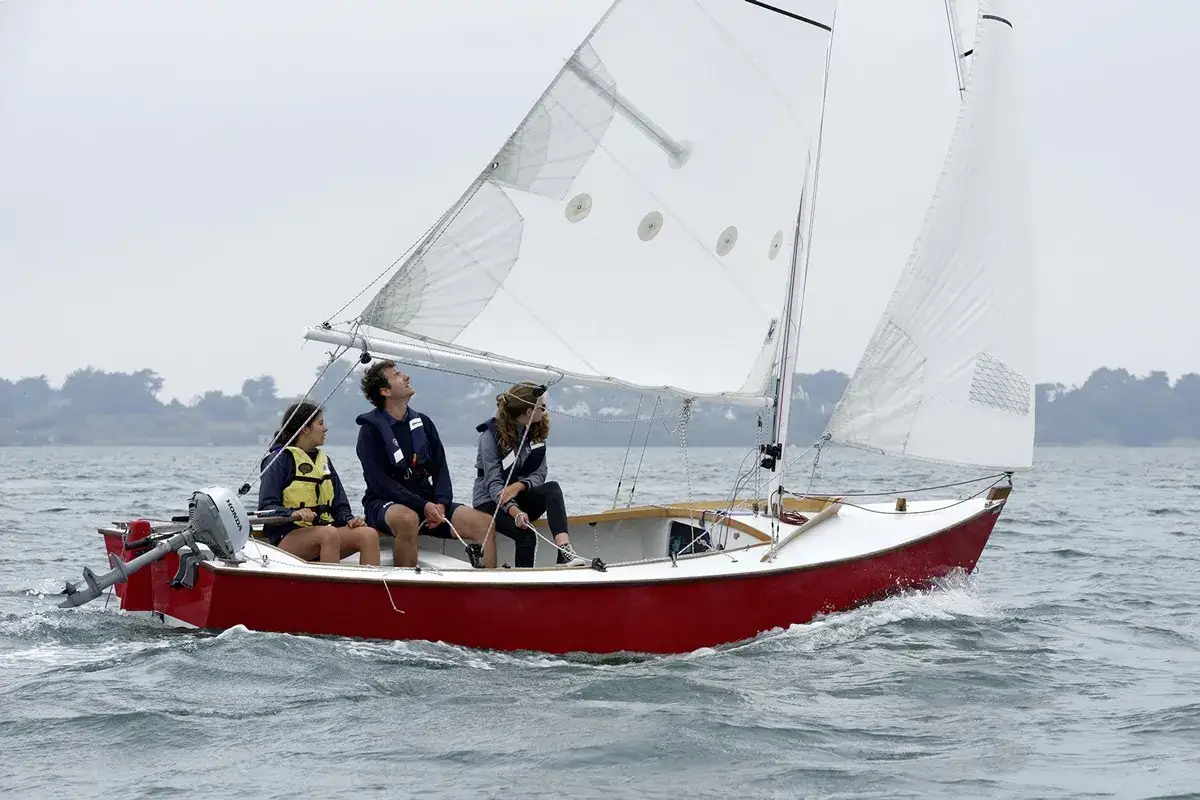 Trois personnes naviguent sur un petit bateau à voile rouge et blanc, profitant d'une journée sur l'eau.