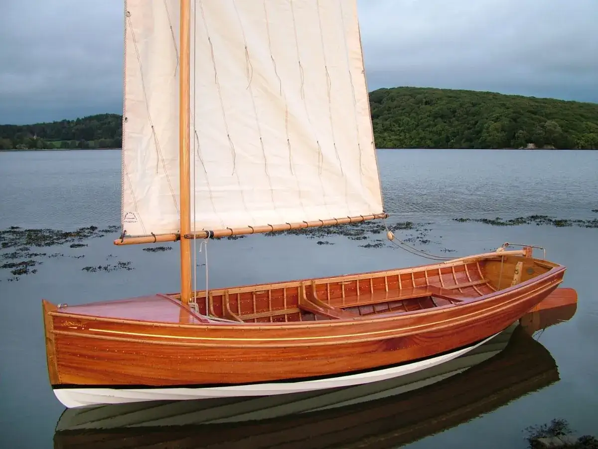 Magnifique folk boat en bois verni, à l'ancre sur une eau calme, avec une voile blanche gonflée par la brise.