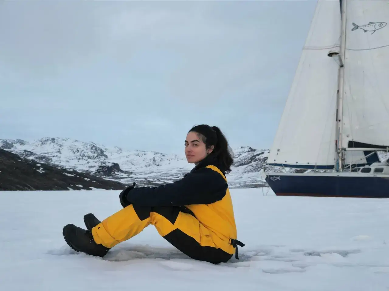 Tamara Klink, aventurière, assise sur la glace, avec son voilier et des montagnes enneigées en arrière-plan.