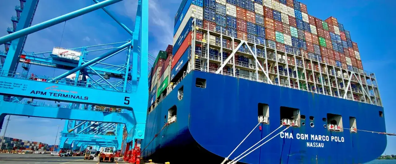 Un immense navire, le CMA CGM Marco Polo, est amarré à quai, prêt à être déchargé par une super porte conteneur bleue d'APM Terminals.