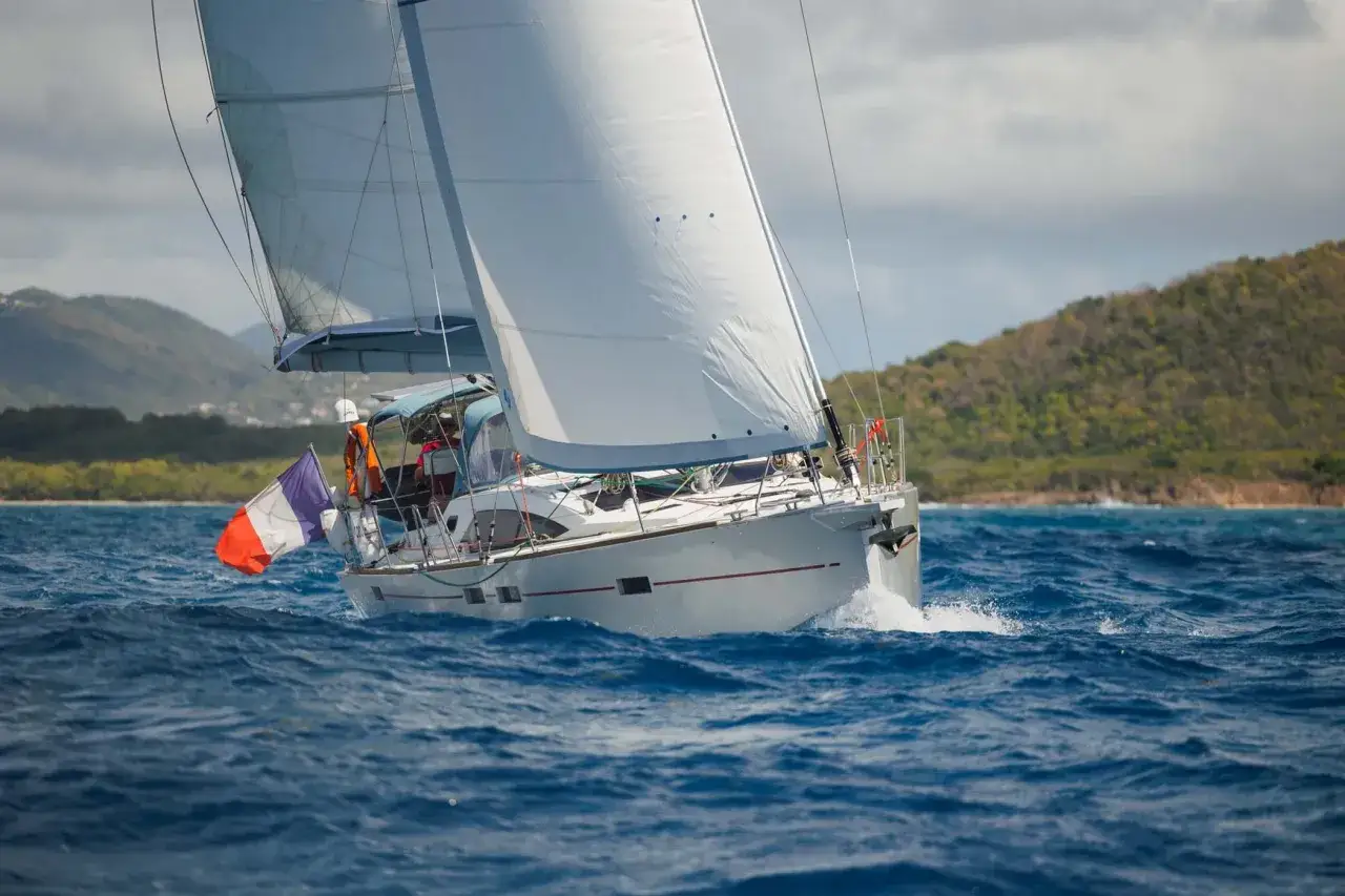 Un voilier blanc navigue sur une mer bleue agitée, pavillon français au vent. L'aventure d'un tour du monde en voilier commence.