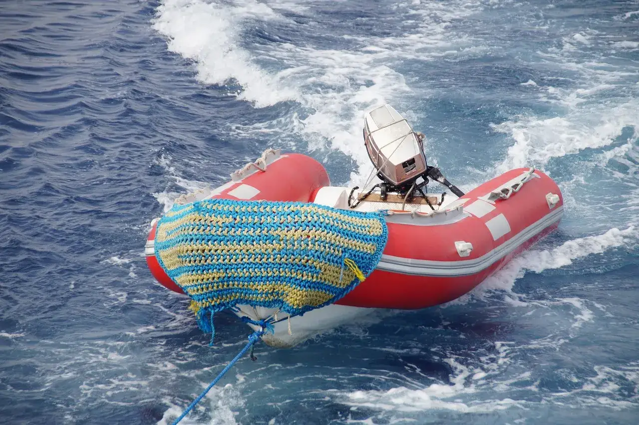 Une annexe rouge, couverte d'une couverture tricotée bleue et jaune, est remorquée sur une mer agitée.