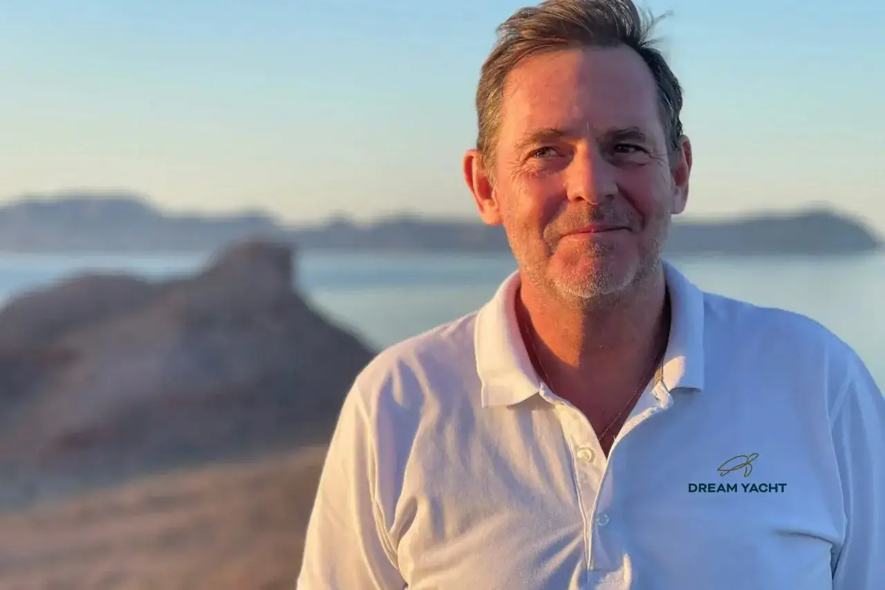 Loïc Bonnet, souriant, porte un polo blanc avec le logo Dream Yacht. En arrière-plan, des montagnes et la mer au coucher du soleil.
