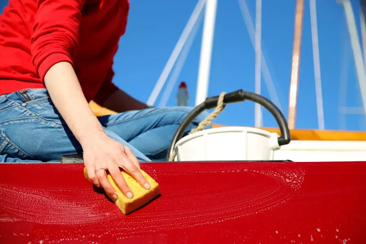 Nettoyage coque bateau vinaigre blanc : une main frotte la coque rouge avec une éponge, sous un ciel bleu.