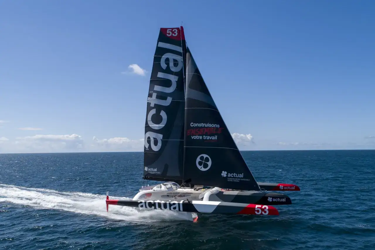 Trimaran Actual, skippé par Yves Le Blevec, fend les vagues bleues sous un ciel clair.