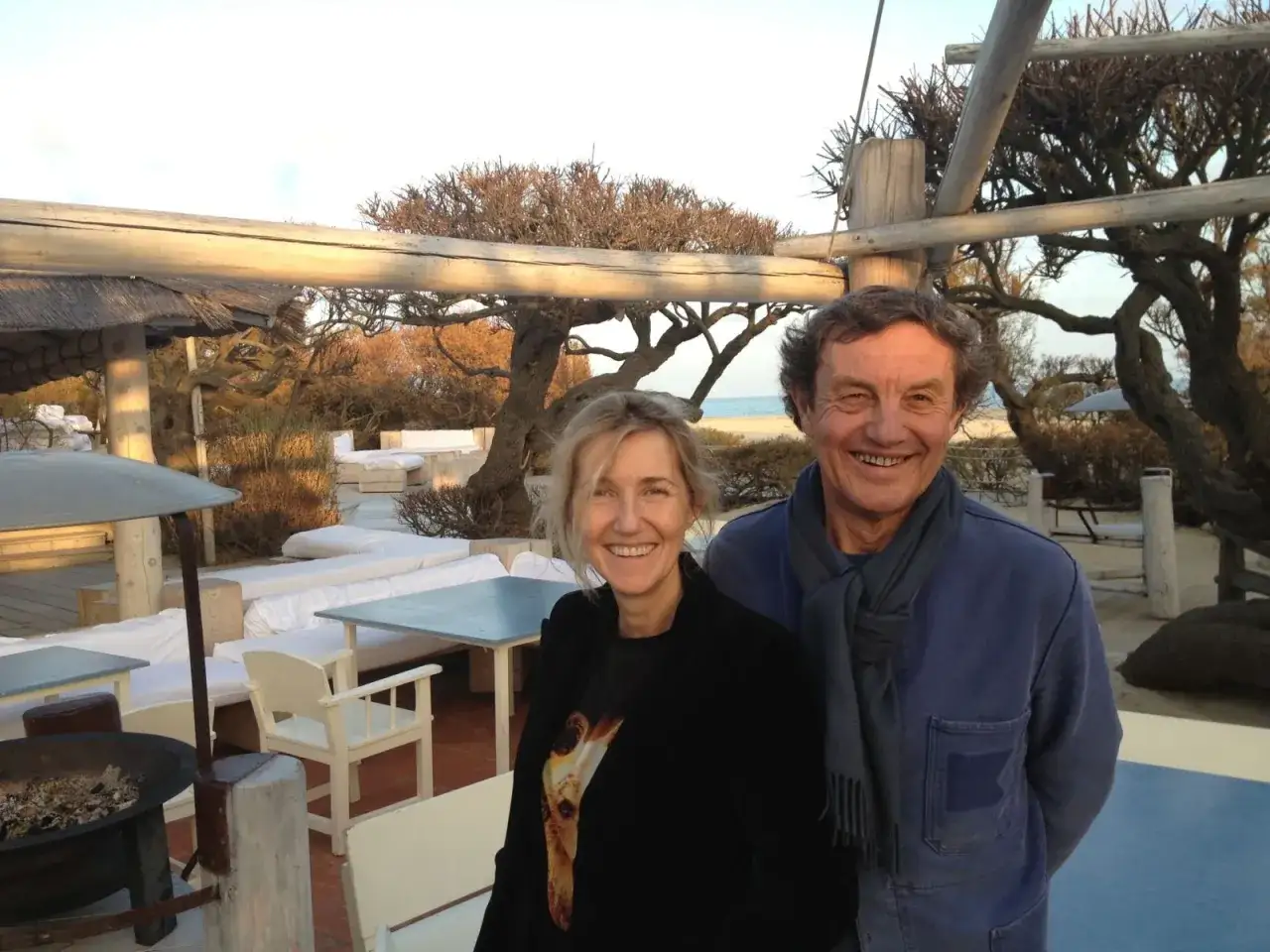 Patrice de Colmont et une femme souriants sur une plage aménagée, avec des arbres tortueux et des sièges blancs en arrière-plan.