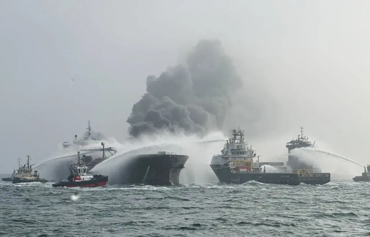 Plusieurs remorqueurs luttent contre un incendie sur un navire, projetant de l'eau sur la structure en feu. La scène évoque une collision bateau dramatique.