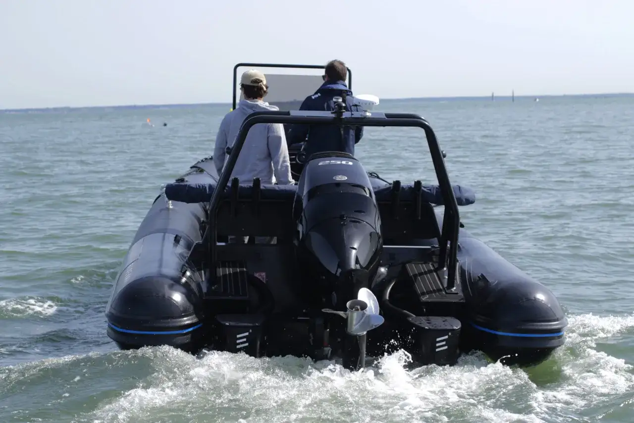 Deux personnes naviguent sur un bateau pneumatique noir, propulsé par un moteur hors-bord. L'eau éclabousse autour du bateau électrique.