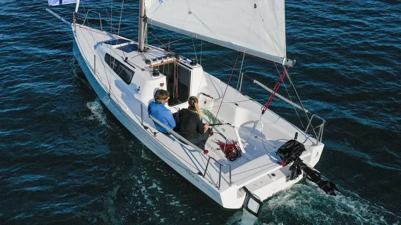 Un voilier First 24 navigue sur une mer calme. Deux personnes sont assises sur le pont, profitant du soleil.