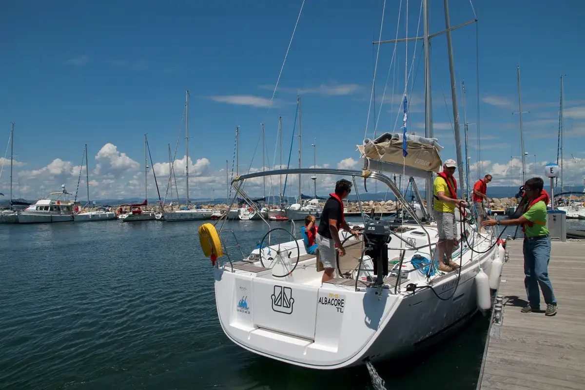 Des hommes préparent un bateau à voile pour le départ. Le soleil brille sur le port, avec d'autres voiliers amarrés.