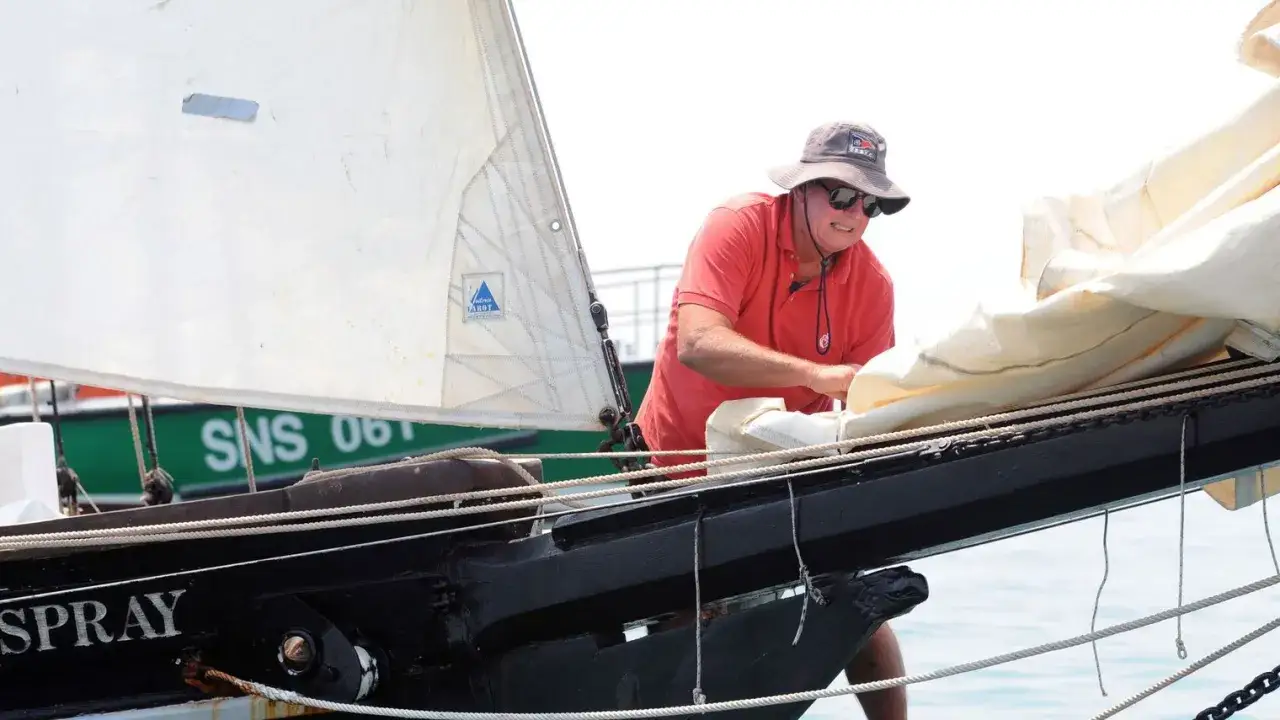 Guy Bernardin, casquette vissée, ajuste les voiles du Spray. Le marin, souriant, est prêt pour la prochaine traversée.