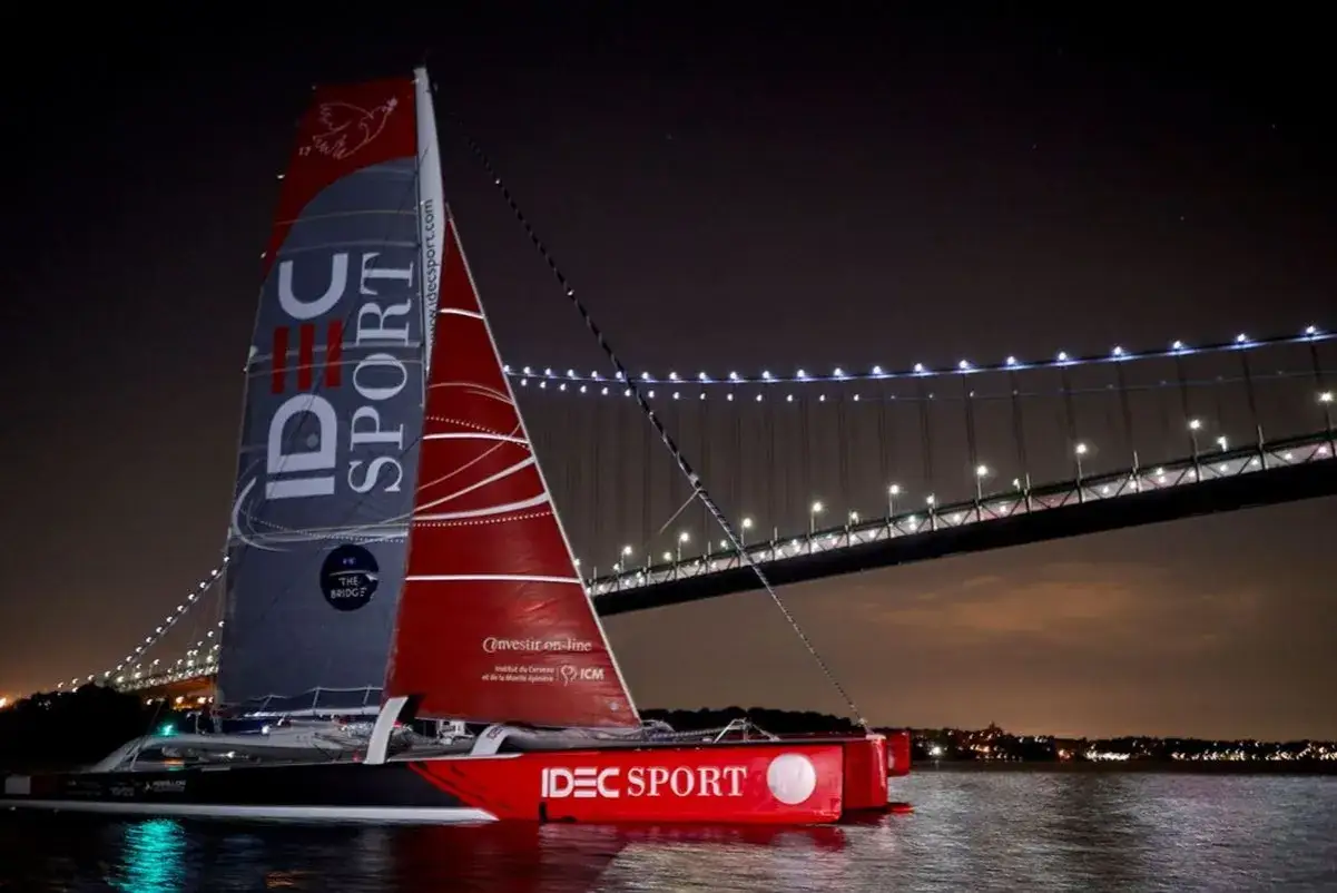 Le trimaran IDEC SPORT, skippé par Francis Joyon, navigue la nuit sous un pont illuminé.