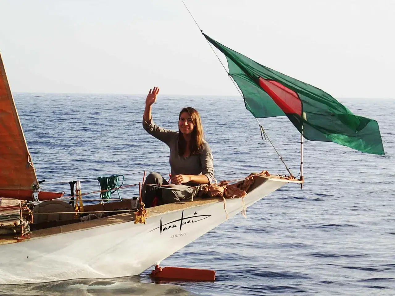 Capucine Trochet salue depuis l'avant d'un voilier, le drapeau du Bangladesh flottant au vent.