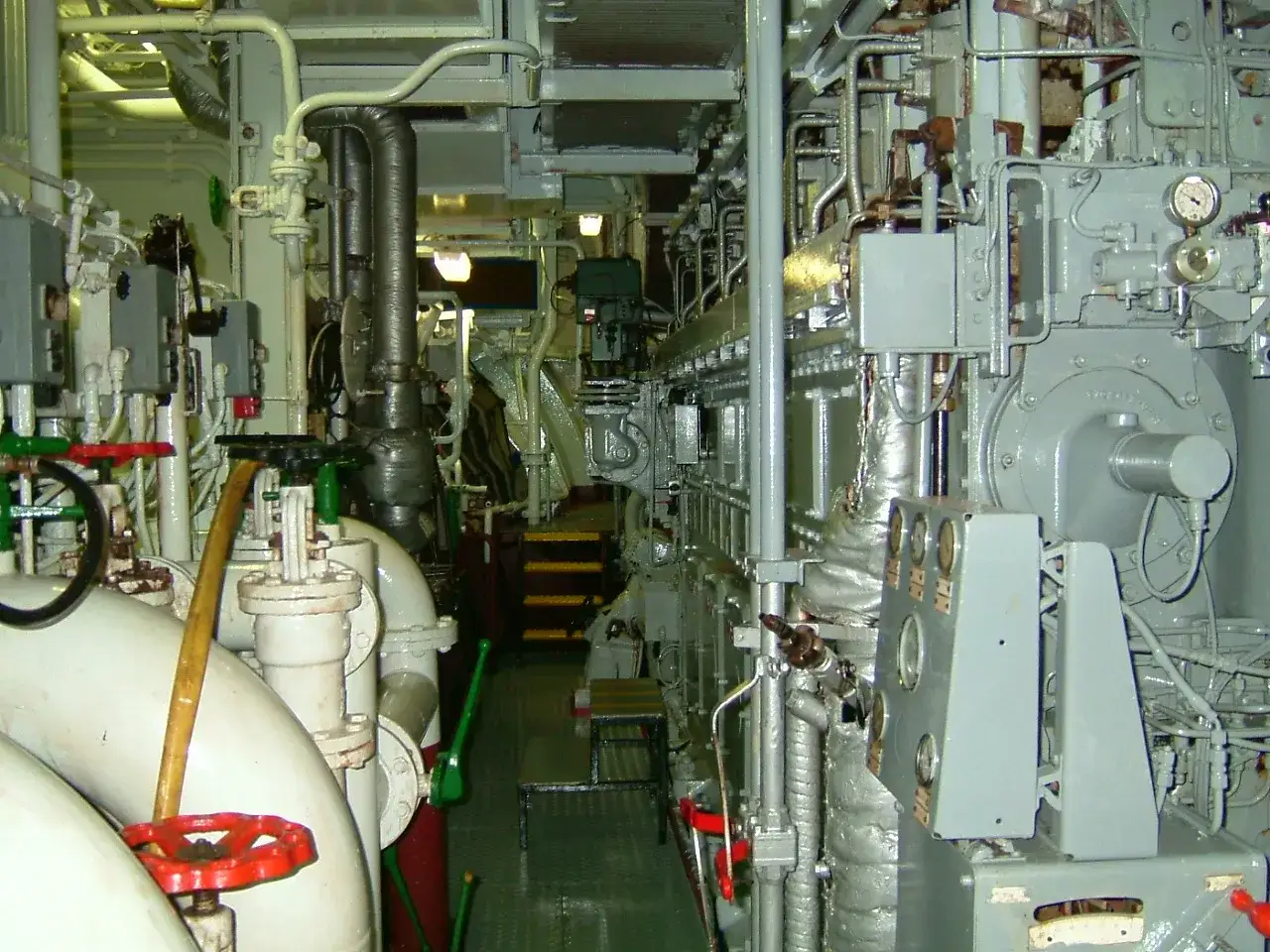 Salle des machines d'un porte-conteneurs, avec un imposant moteur diesel, tuyauterie et vannes.