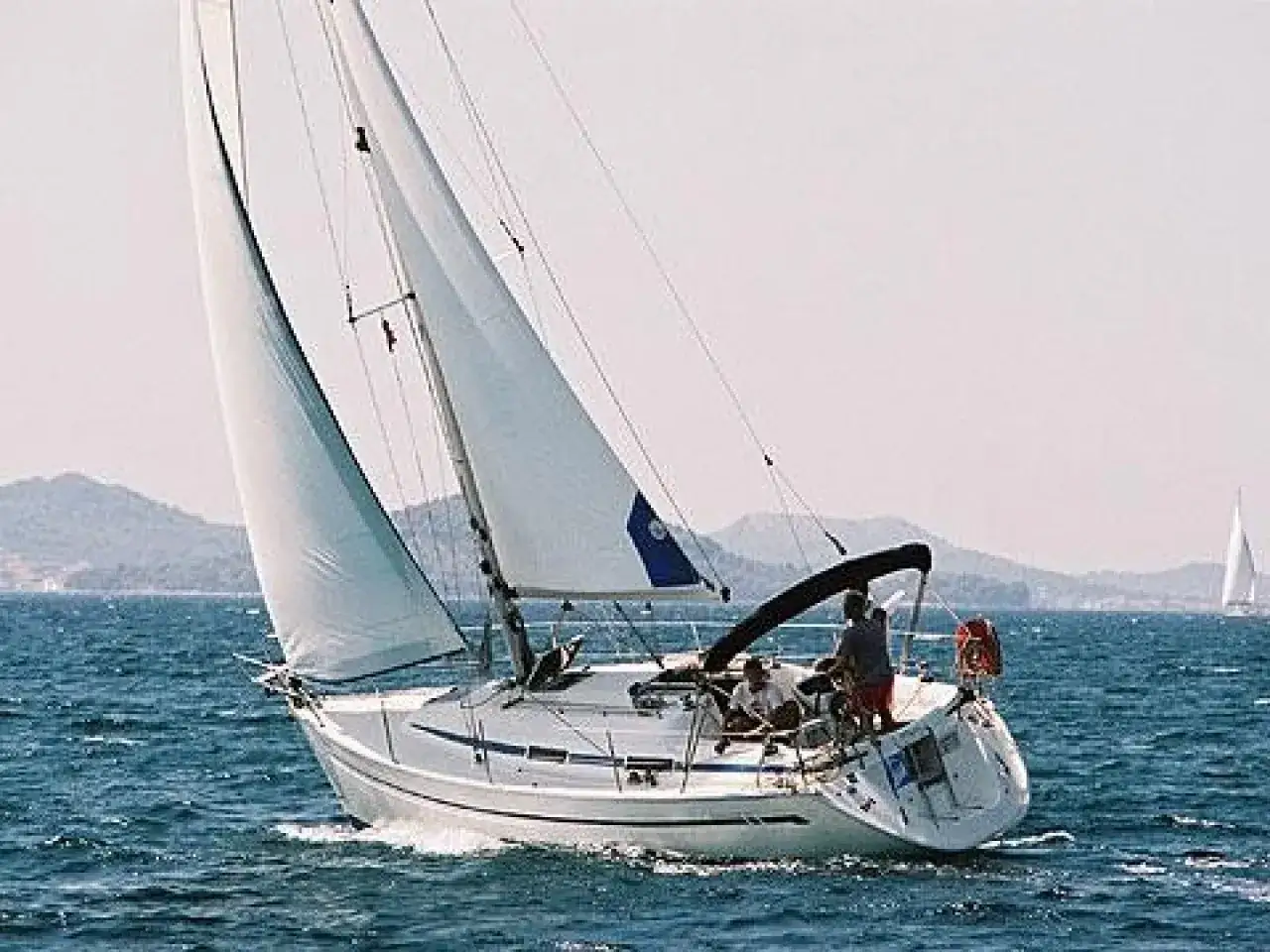 Bavaria 32 avis : un voilier blanc navigue sur une mer bleue, voiles gonflées, avec des collines en arrière-plan.