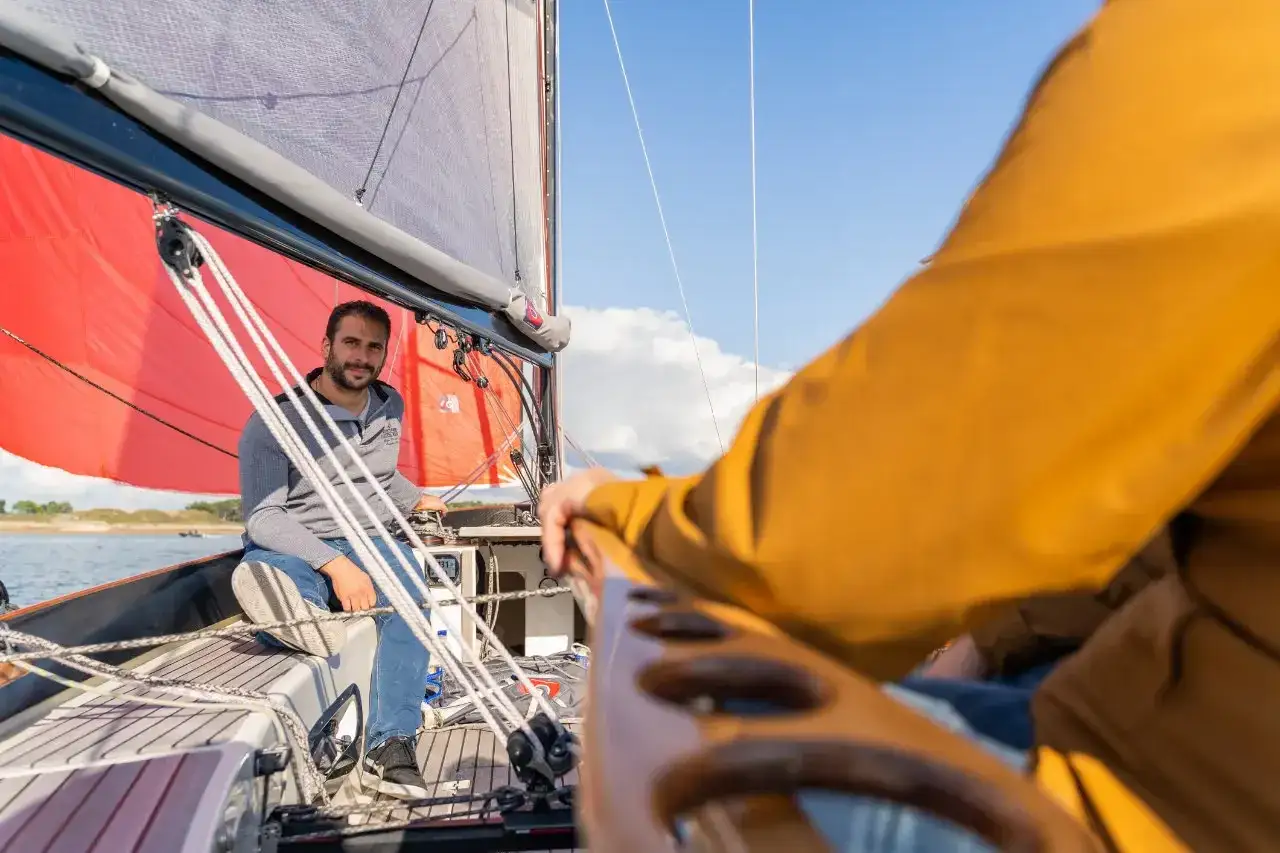 Un homme souriant sur un voilier, une voile rouge déployée. On dirait qu'il nous montre comment naissent les bateaux, en pleine mer.