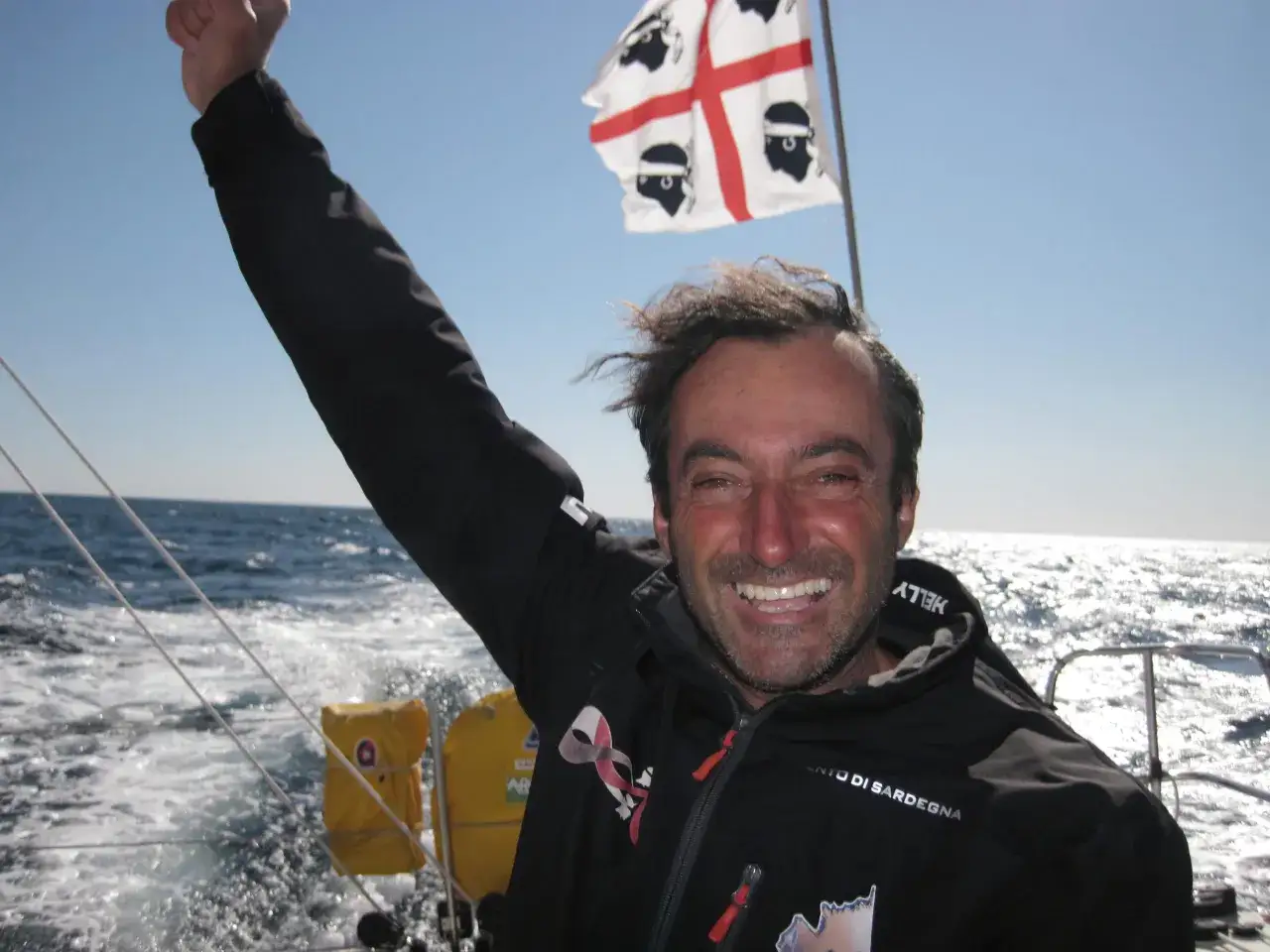 Giancarlo Pedote, fier de sa victoire, lève le poing sur son bateau, le drapeau de la Sardaigne flottant au vent.