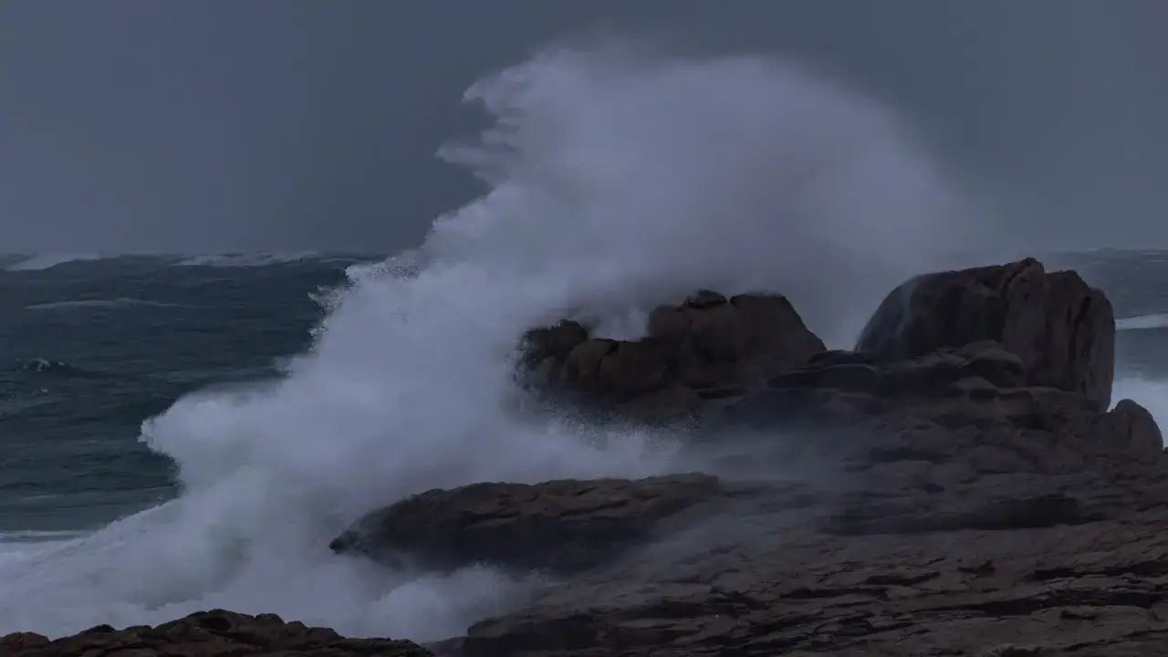 Vague d'écume s'écrasant sur des rochers sombres, comme un bms bateau en pleine tempête. La mer déchaînée et le ciel menaçant créent une atmosphère dramatique.