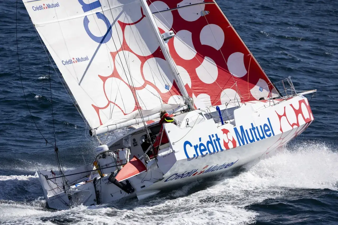 Le voilier Crédit Mutuel, skippé par Corentin Douguet, fend les vagues bleues, ses voiles ornées de cercles rouges et blancs.