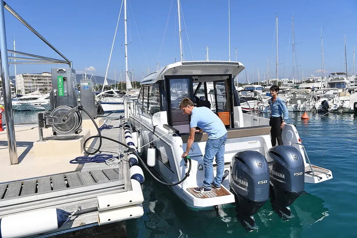 Un homme fait le plein d'un bateau avec un moteur hors-bord essence 95 ou 98. La scène se déroule dans un port animé.