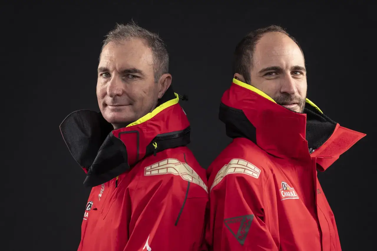 Deux marins, dont Morgan Lagravière, portent des vestes rouges de voile, prêts pour l'aventure en mer.