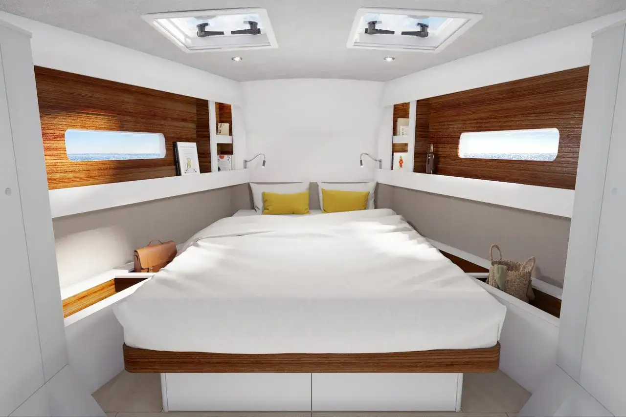 Cabine confortable d'un voilier habitable à l'année, avec un grand lit blanc, des hublots donnant sur la mer et des puits de lumière.