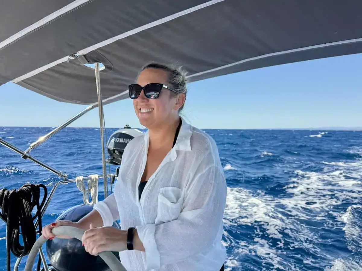 Une femme souriante tient la barre d'un voilier, le soleil brille sur la mer bleue. Un moment de pur bonheur pour ce récit de voyage en voilier.
