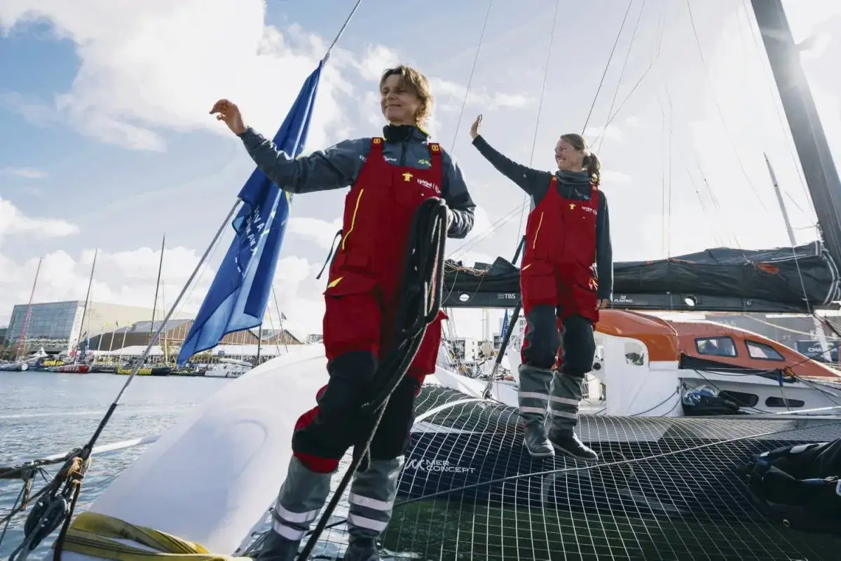 Anne Claire Le Berre et une autre navigatrice saluent depuis un catamaran, prêtes pour une course.