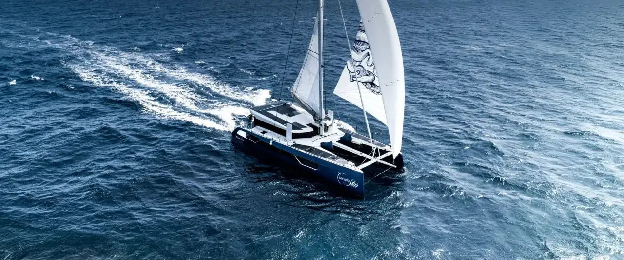 Un catamaran bleu navigue sur une mer agitée, ses voiles blanches gonflées par le vent. Ce navire est le meilleur catamaran hauturier, prêt pour l'aventure.