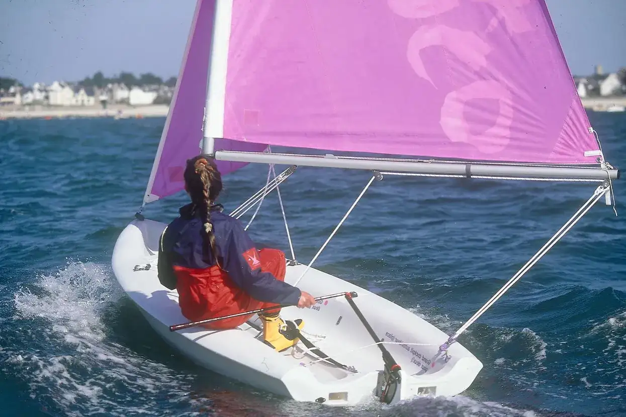 Jeune fille aux cheveux tressés, profitant du plaisir de faire la voile sur un petit bateau blanc, sous une voile rose vif.