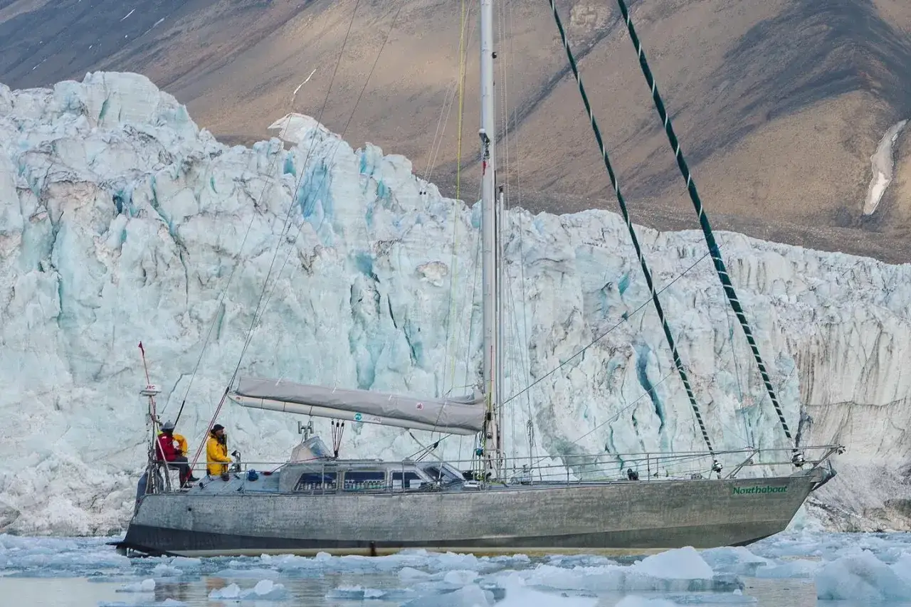 Un voilier, symbole de la persévérance, navigue près d'un glacier imposant. Deux personnes observent le paysage glacial.