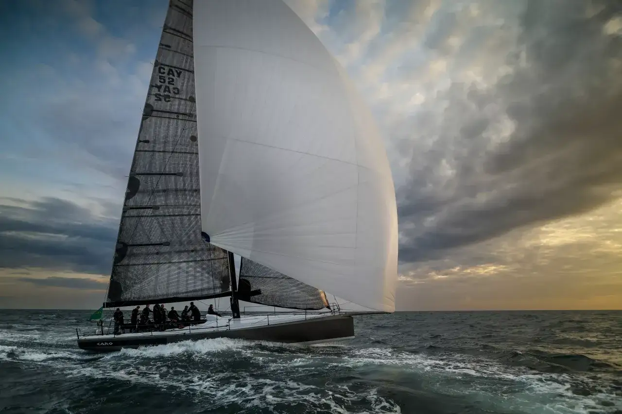 Un voilier de course fend les vagues sous un ciel dramatique, son spi symétrique blanc gonflé par le vent. L'équipage est concentré sur la manœuvre.