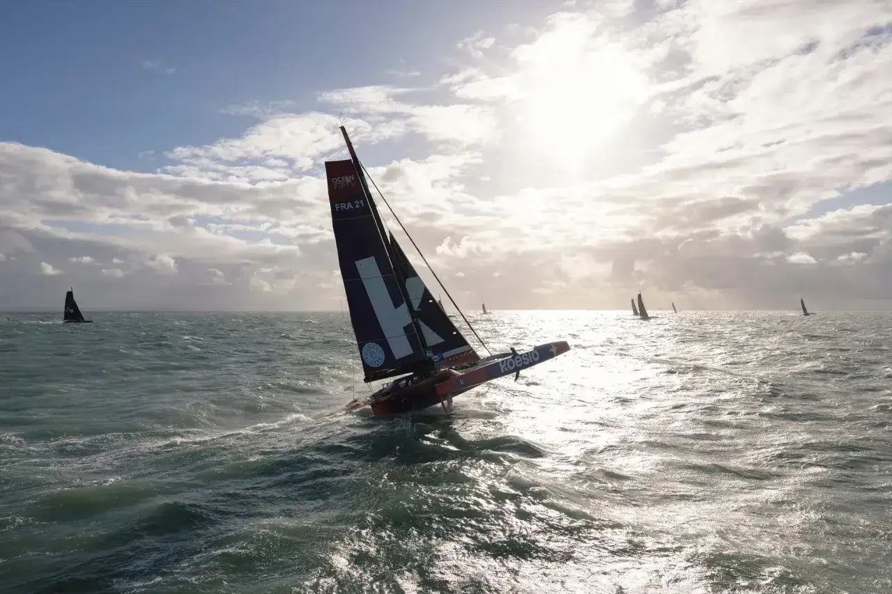 Le trimaran Koesio de Matthieu Perraut fend les vagues sous un ciel nuageux, participant à une course avec d'autres voiliers.