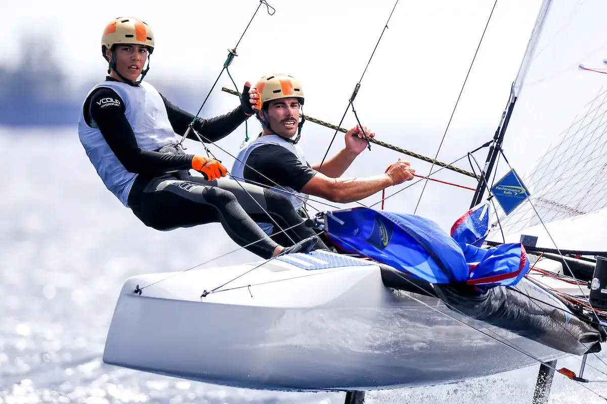 Deux jeunes hommes en équipement de voile, dont Lou Berthomieu, naviguent sur un catamaran, le vent gonflant leurs voiles.