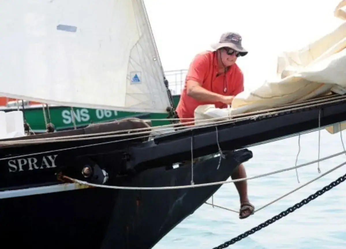Guy Bernardin, casquette et lunettes, ajuste une voile sur le pont du voilier