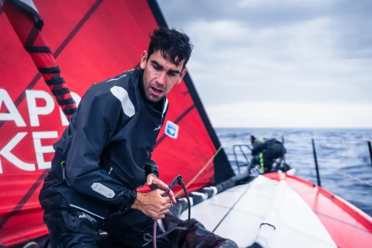 Gaston Morvan, concentré, manœuvre une voile rouge sur un voilier en pleine mer. L'océan est agité sous un ciel couvert.