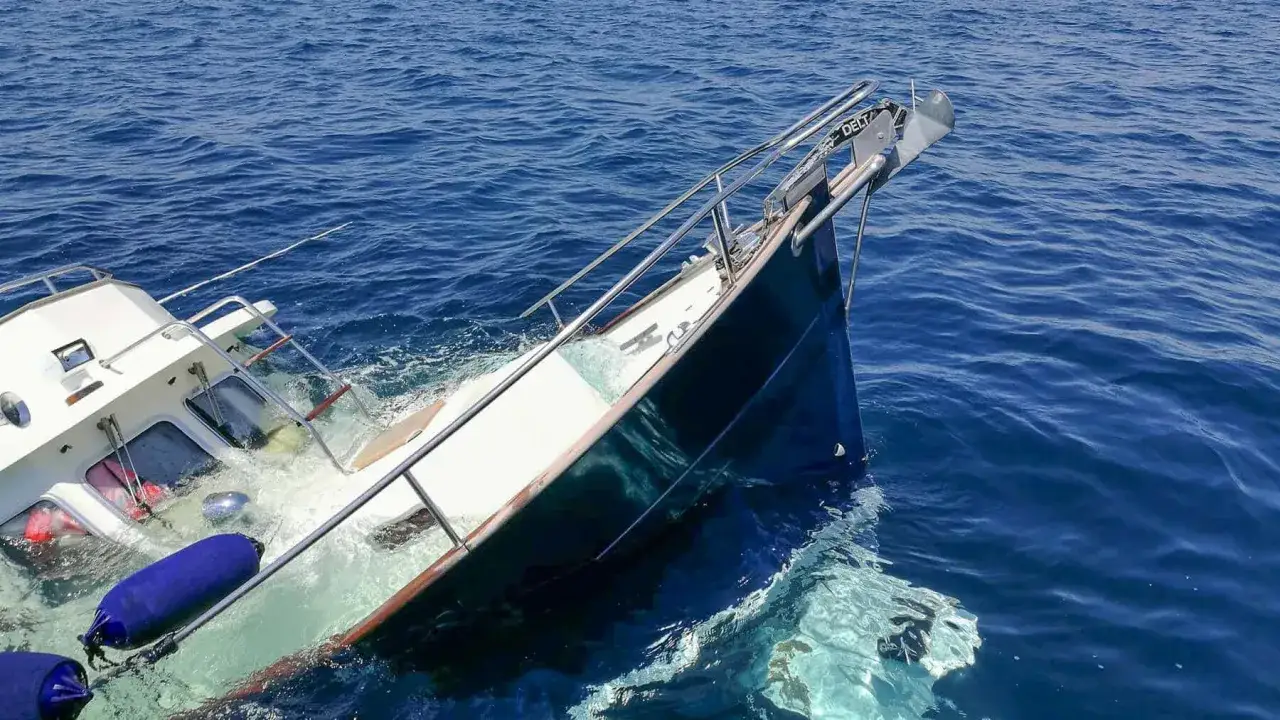 Un bateau coule, sa proue hors de l'eau, sur une voie d'eau. Des pare-battages bleus flottent à côté.