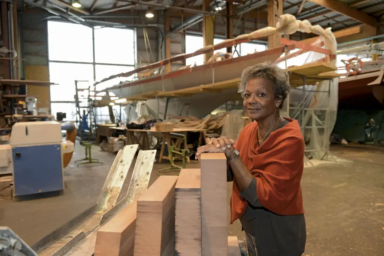 Jacqueline Tabarly, souriante, dans un atelier de construction navale, devant des blocs de bois et un voilier en cours de fabrication.