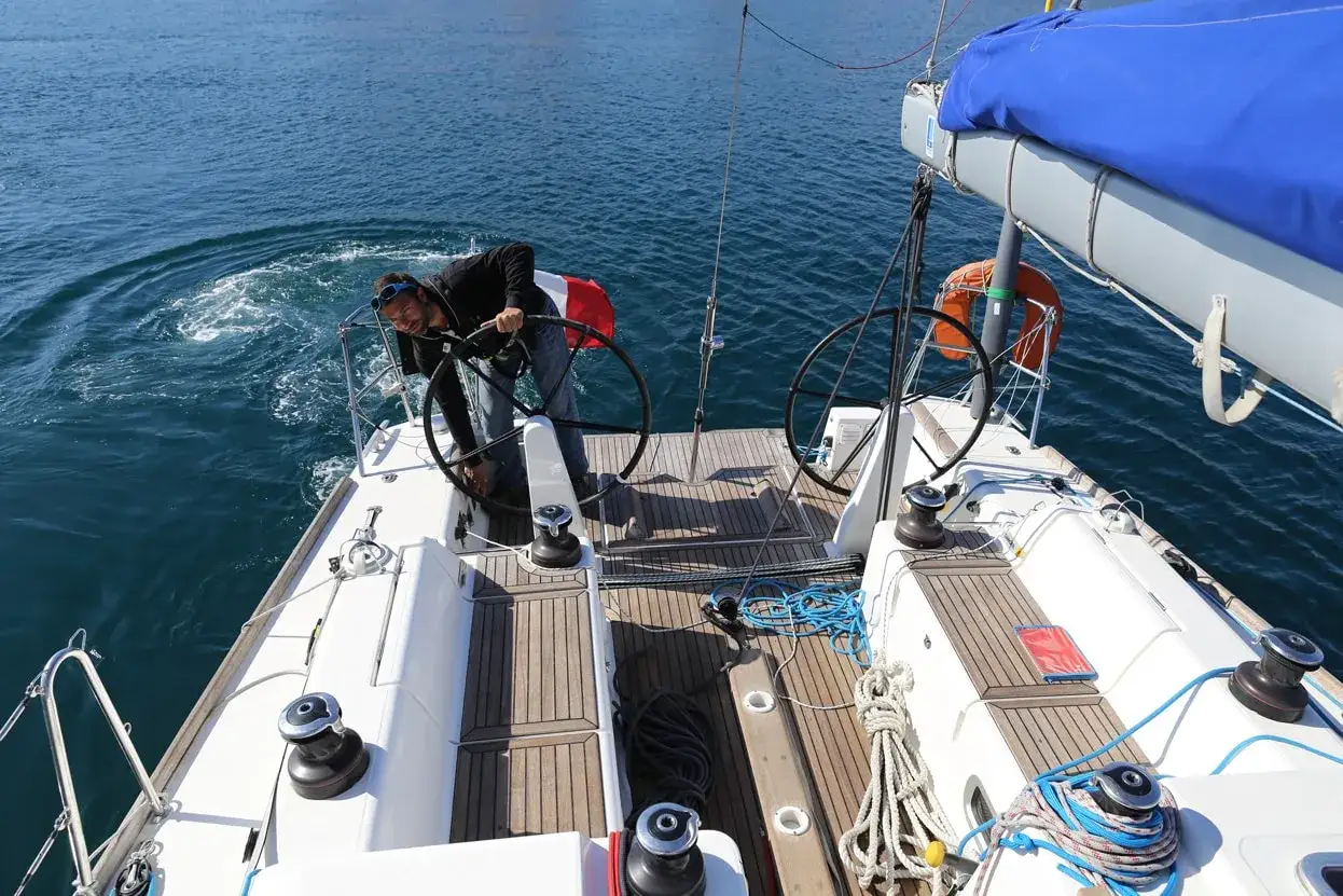 Le chef de bord bateau manœuvre la barre, créant des remous dans l'eau bleue. Le soleil brille sur le pont en bois.