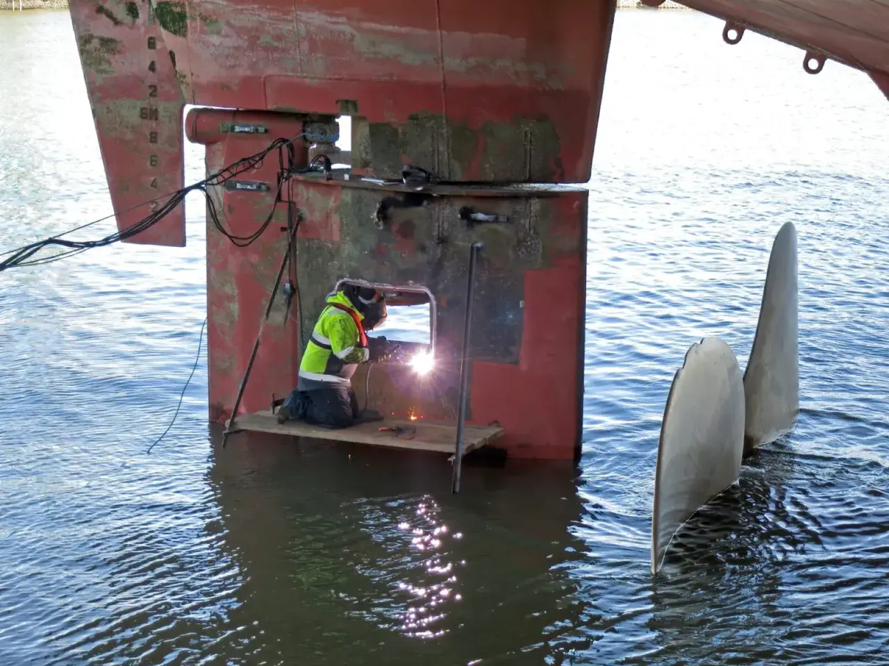 Un soudeur travaille sur la coque d'un safran voilier, entouré par l'eau.