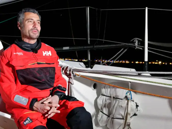 Giancarlo Pedote, skipper en tenue de voile rouge, sur un bateau la nuit, avec des lumières de ville en arrière-plan.