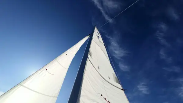 Voiles blanches gonflées par le vent, le bateau semble s'affaler sur l'eau bleue sous un ciel clair.
